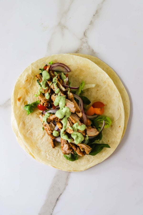 An open flour tortilla topped with grilled chicken, mixed greens, sliced red onions, tomatoes, and a green sauce, on a white marble surface.