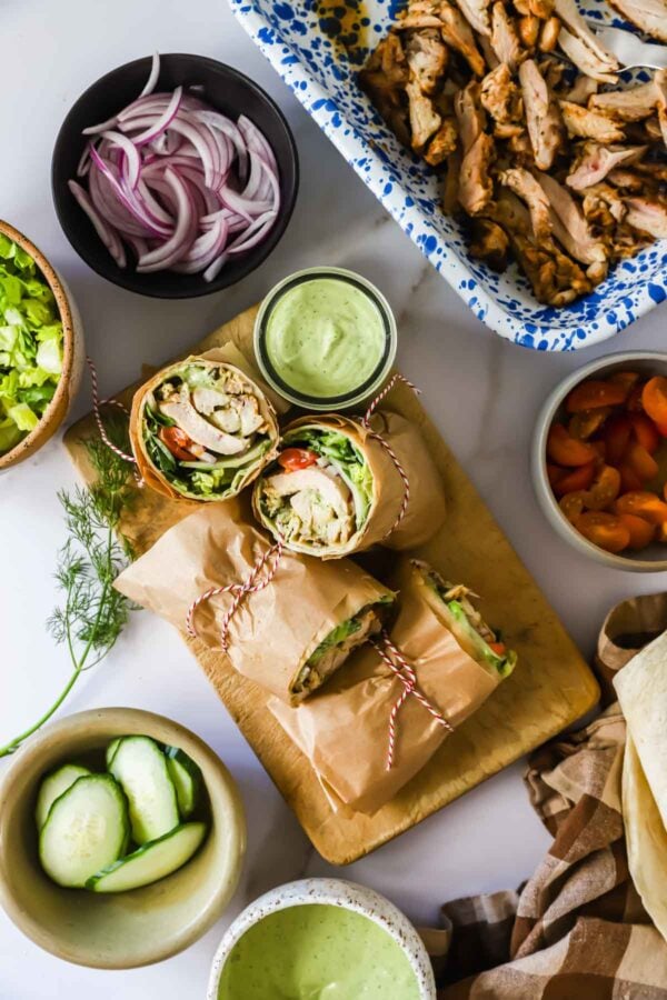 Overhead view of chicken wraps on a board, surrounded by bowls of sliced cucumber, red onion, greens, sauce, cooked chicken, and sliced carrots.
