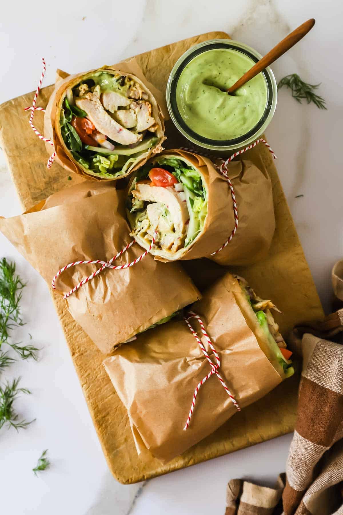 Three chicken wraps tied with red and white string sit on a wooden board next to a jar of green sauce with a wooden spoon, garnished with fresh dill.