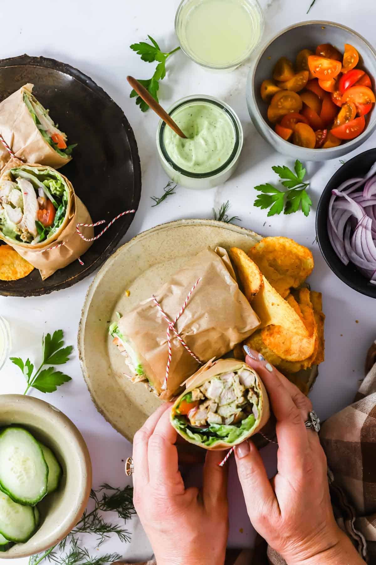 A person holds a wrapped chicken wrap over a plate with chips. Other plates contain another wrap, sliced tomatoes, red onions, cucumbers, sauce, and fresh herbs.