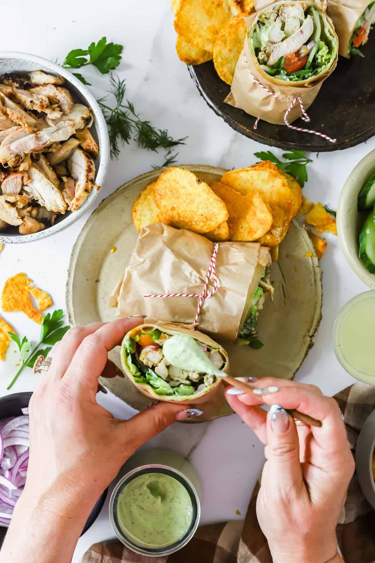 Overhead view of a person holding a wrapped chicken wrap on a plate with chips; additional chicken, wraps, and drinks are visible on the table.