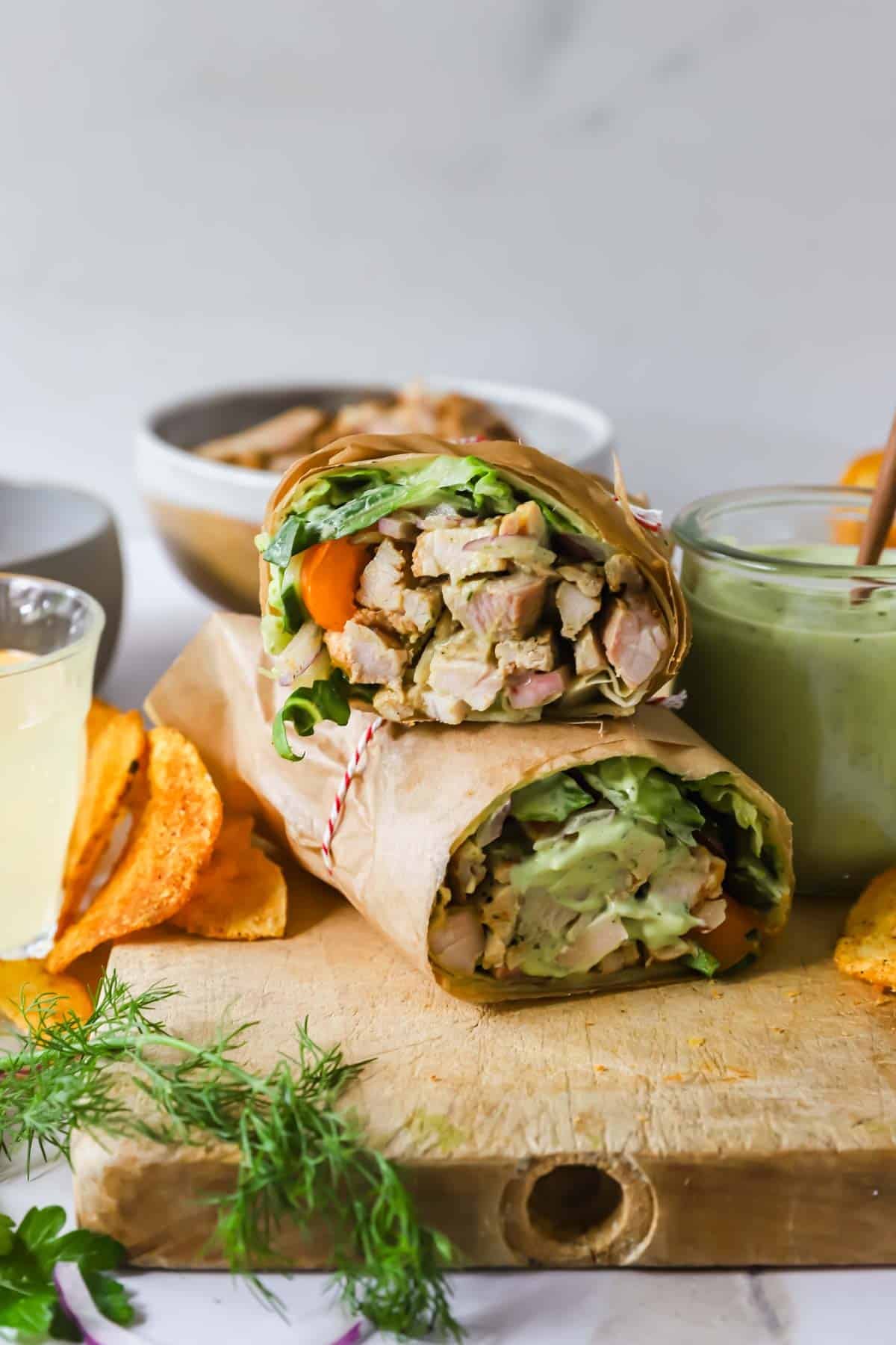 Two chicken wraps with lettuce and sauce are stacked on a wooden board, accompanied by potato chips, a jar of green sauce, and fresh herbs.