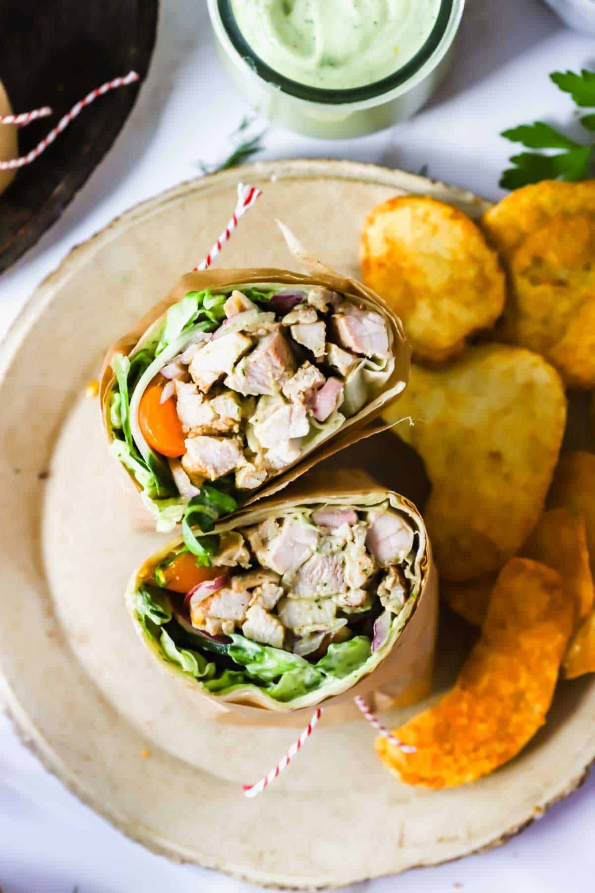 A plate with a cut chicken wrap filled with lettuce and tomatoes, served alongside hash browns and orange potato chips.