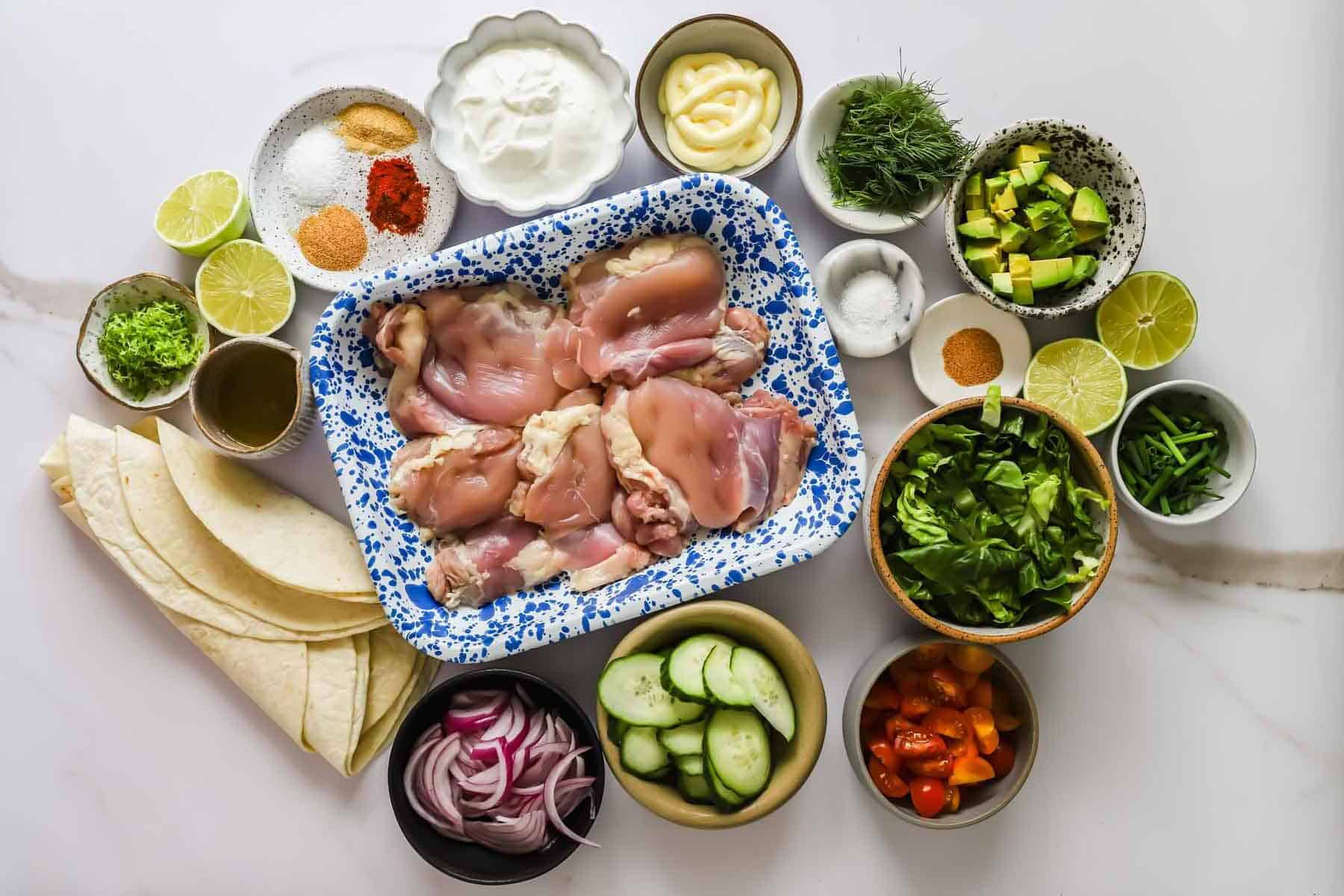 A top view of raw chicken thighs on a tray, surrounded by small bowls of fresh vegetables, herbs, seasonings, sauces, lime halves, and tortillas on a white surface.