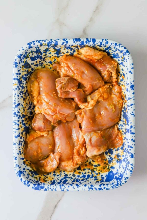 Raw marinated chicken thighs arranged in a blue and white speckled tray on a white marble surface.