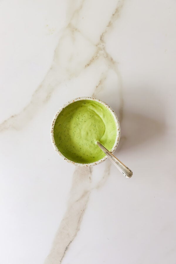 A small bowl filled with creamy green sauce or dressing, with a spoon resting inside, placed on a white marble surface.