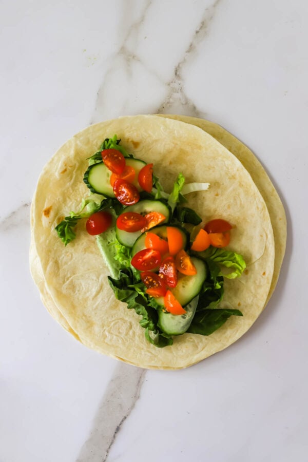 Two flour tortillas topped with fresh lettuce, sliced cucumbers, and chopped cherry tomatoes on a white marble surface.