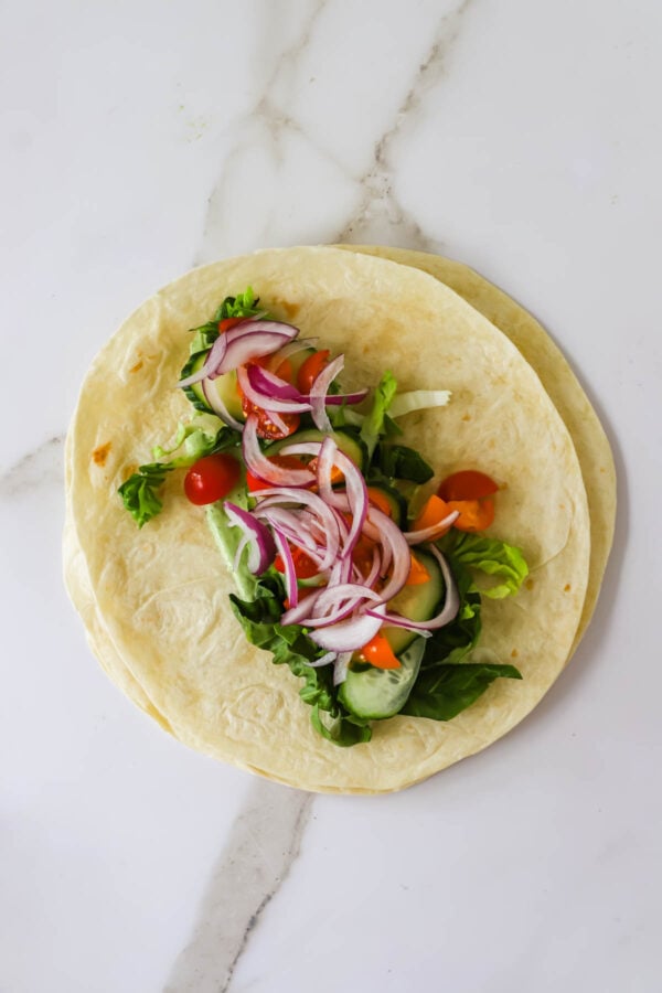 Two flour tortillas topped with mixed greens, sliced cucumbers, cherry tomatoes, and red onion on a white marble surface.