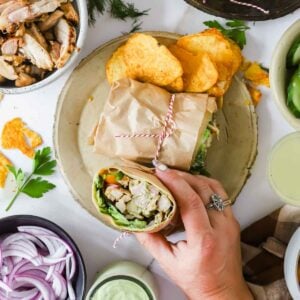 A hand holds a wrap filled with chicken, lettuce, and vegetables on a plate with more wrap halves and potato chips; bowls of onions, sauce, and chicken surround the plate.