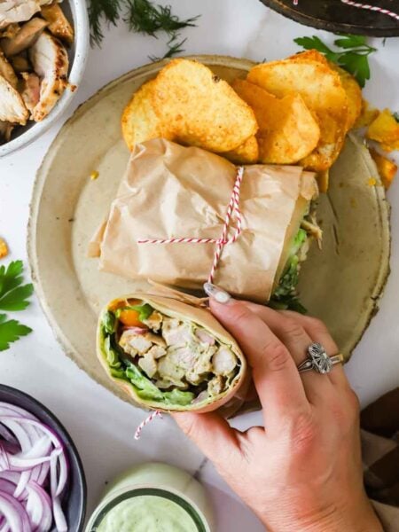 A hand holds a wrap filled with chicken, lettuce, and vegetables on a plate with more wrap halves and potato chips; bowls of onions, sauce, and chicken surround the plate.