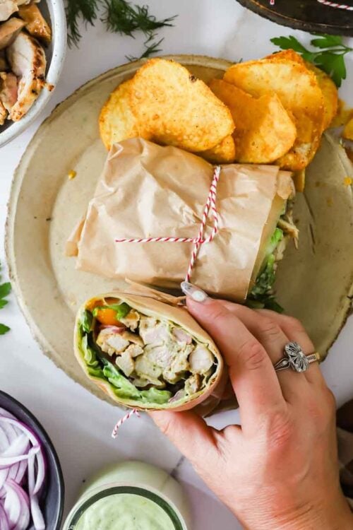 A hand holds a wrap filled with chicken, lettuce, and vegetables on a plate with more wrap halves and potato chips; bowls of onions, sauce, and chicken surround the plate.