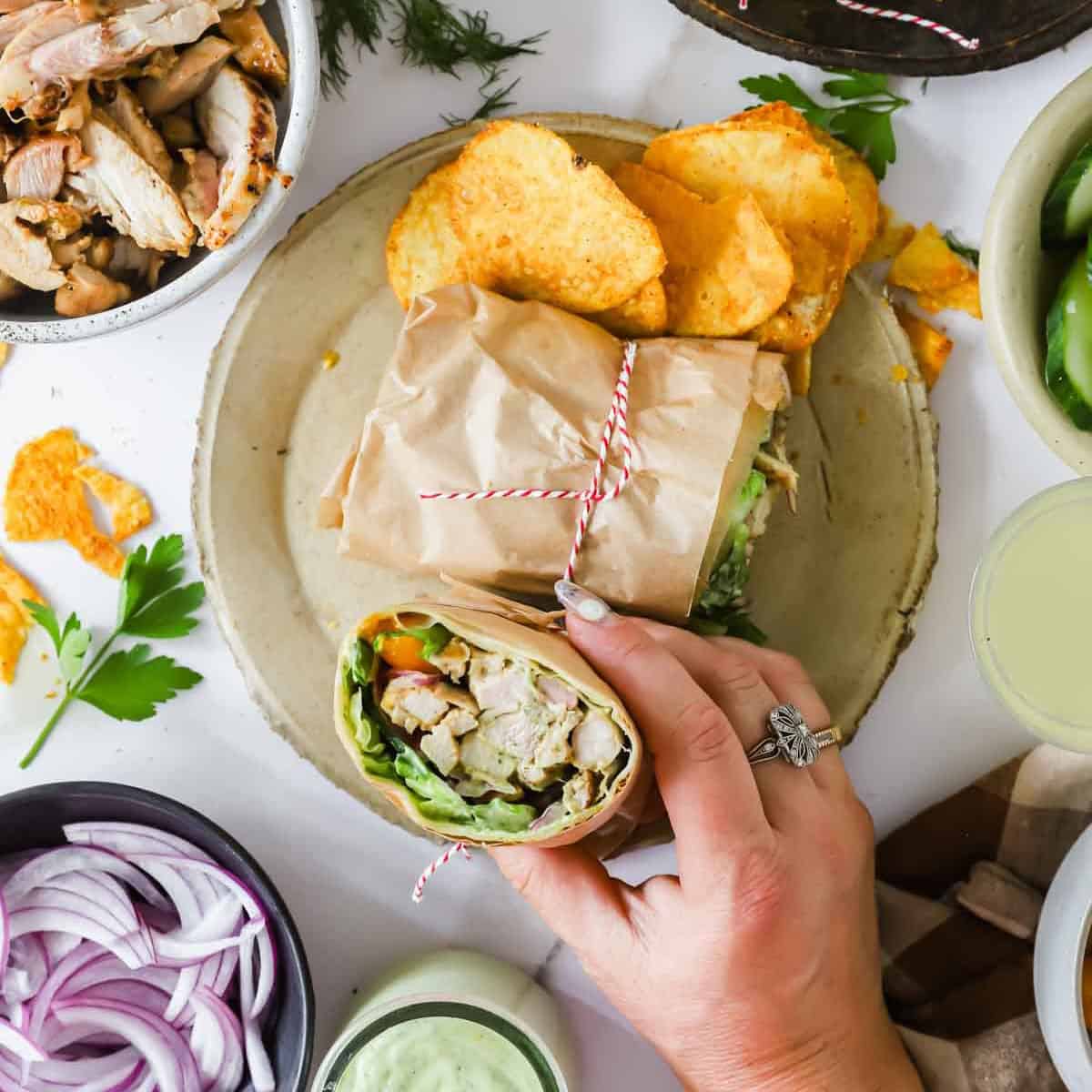 A hand holds a wrap filled with chicken, lettuce, and vegetables on a plate with more wrap halves and potato chips; bowls of onions, sauce, and chicken surround the plate.