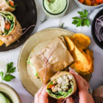 Hands holding a grilled chicken wrap on a plate with chips, surrounded by sauces, vegetables, and garnish; text overlay reads "Easy Grilled Chicken Wraps.