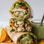 Three grilled chicken wraps stacked on a cutting board, sliced open to show lettuce, tomato, and sauce, with chips and a jar of green dip on the side.