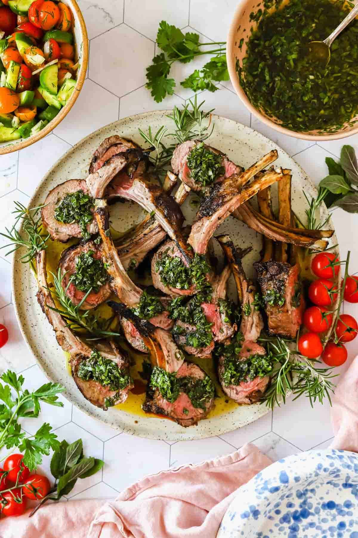 A plate of grilled lamb chops topped with green herb sauce, garnished with sprigs of rosemary and cherry tomatoes, with salad and sauce bowls on the side.