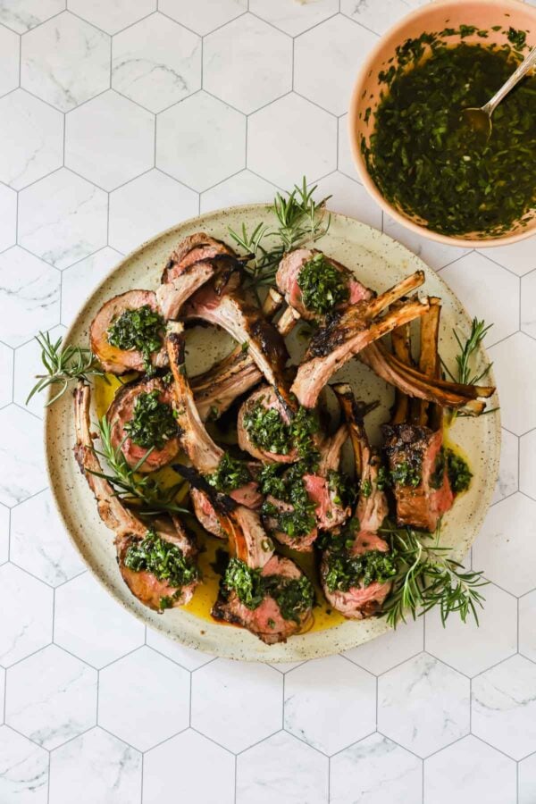 A plate of grilled lamb chops garnished with green herb sauce, served with extra sauce in a bowl on a white hexagon-tiled surface.