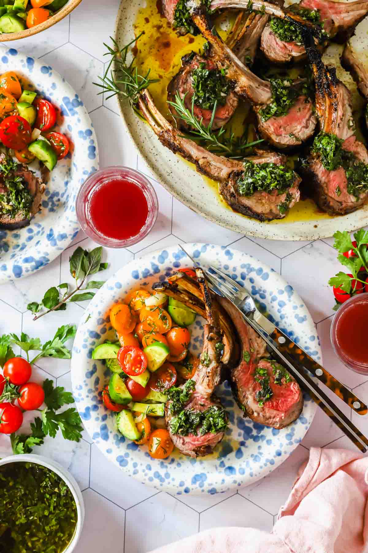 Plates of grilled lamb chops with green herb sauce, served with tomato and cucumber salad, next to glasses of red drink on a white table.