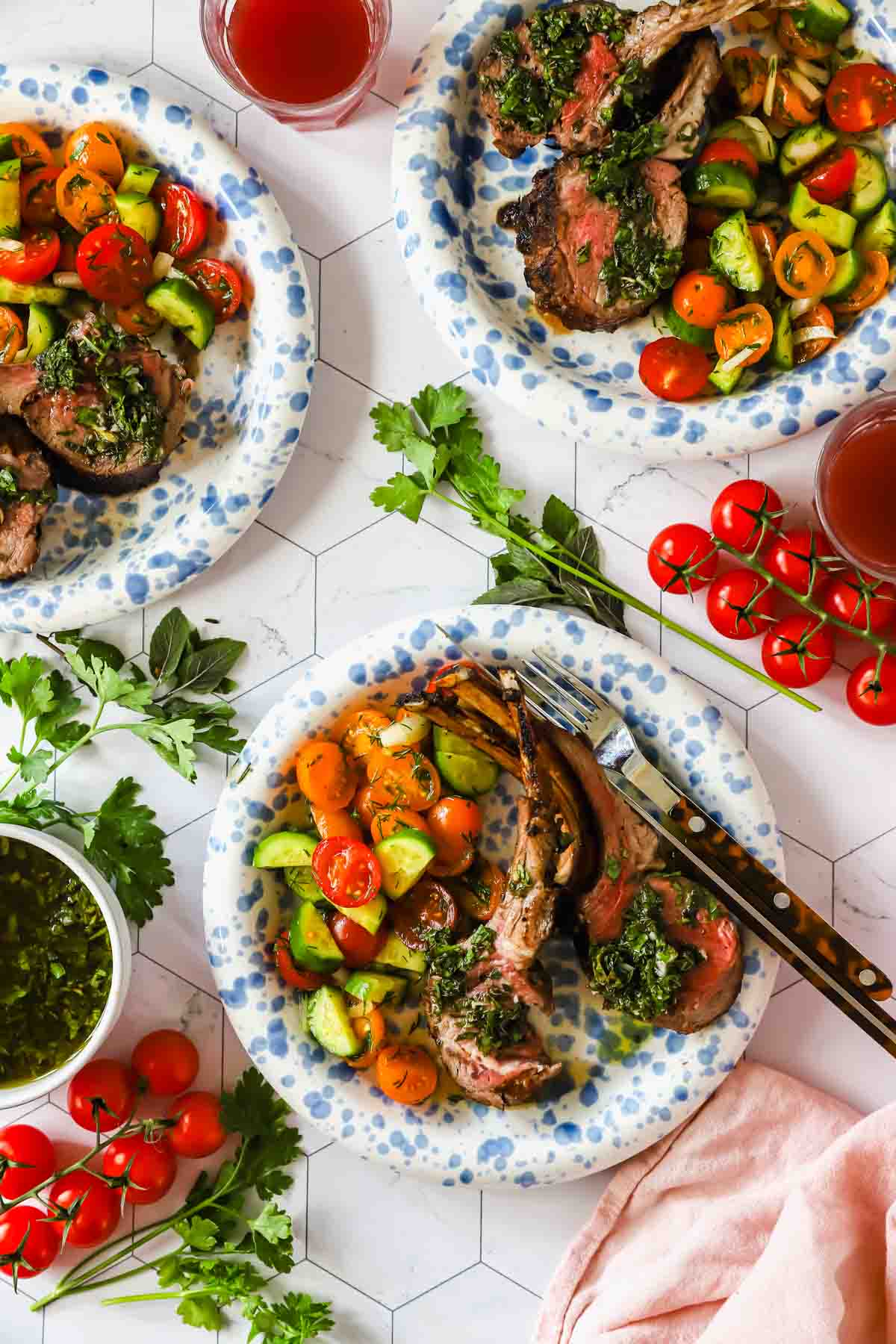 Three plates with grilled lamb chops topped with green sauce, served with a tomato and cucumber salad. Fresh herbs, cherry tomatoes, and drinks are on the table.