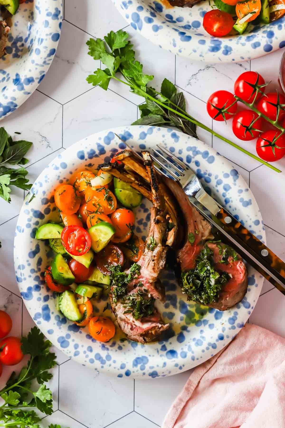 A plate with two lamb chops topped with green herb sauce, served with a side of cucumber and cherry tomato salad. A fork and knife rest on the plate.