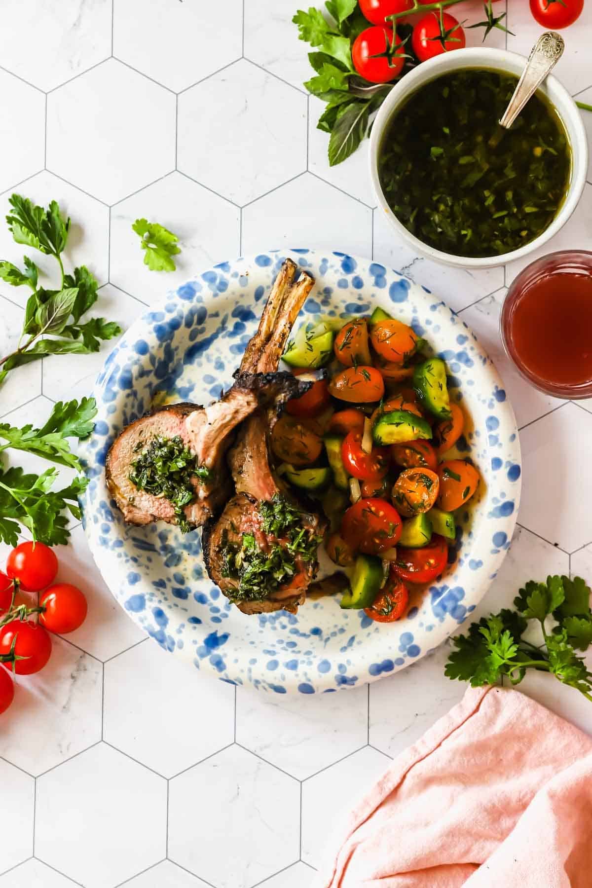 A plate with two grilled lamb chops topped with green herb sauce, served with a tomato and cucumber salad; garnishes and sauce bowl nearby on a white patterned surface.