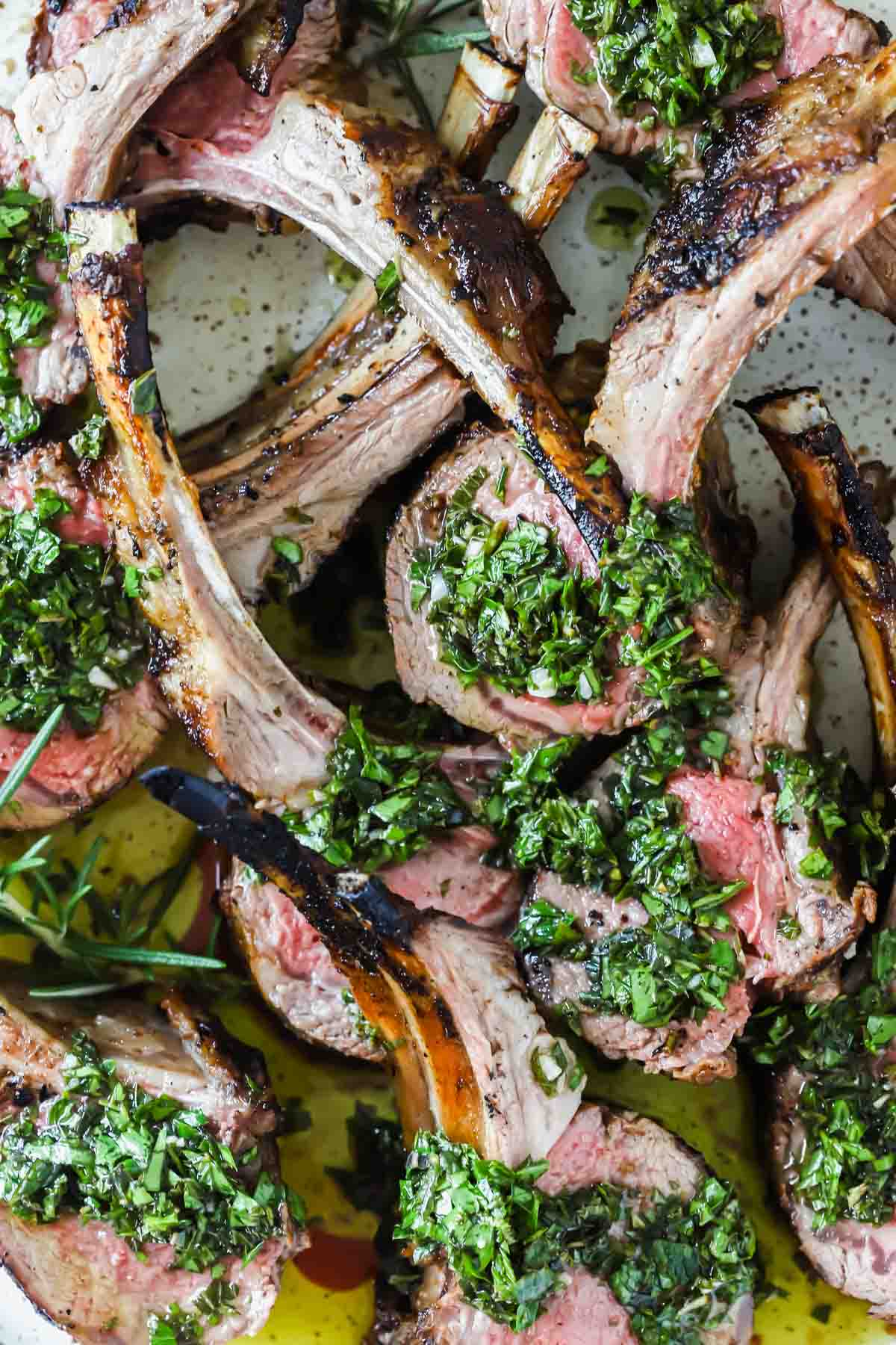Grilled lamb chops arranged closely together, topped with a green herb sauce and garnished with herbs, served on a white plate.