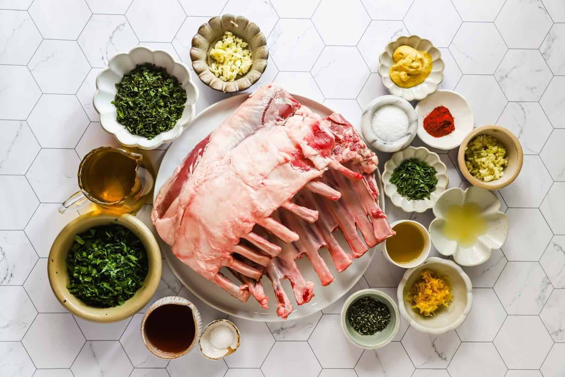 Two raw racks of lamb on a plate, surrounded by small bowls containing chopped herbs, spices, mustard, minced garlic, lemon zest, salt, oil, and other seasonings on a white surface.