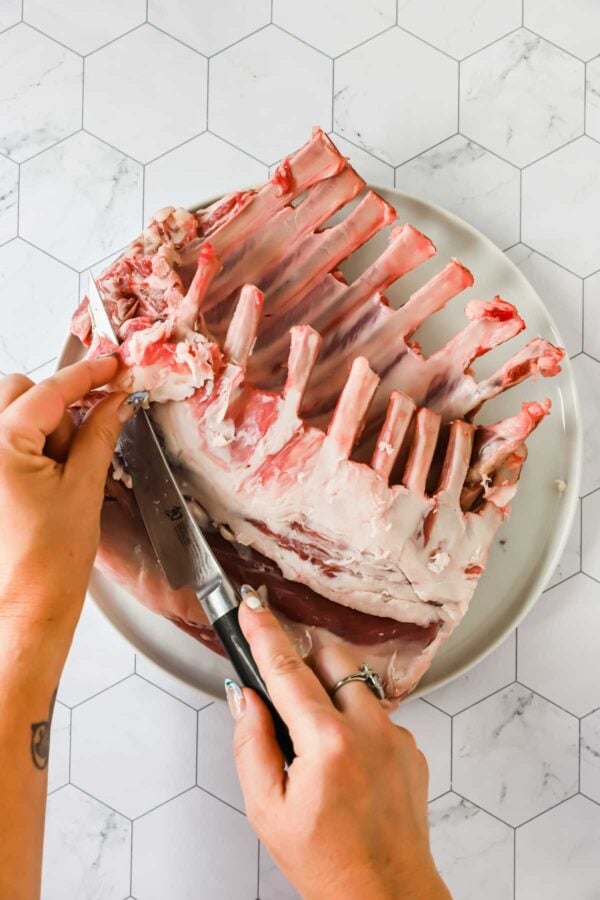Person trimming excess fat from a raw rack of lamb with a knife on a white plate, placed on a white hexagonal tile surface.