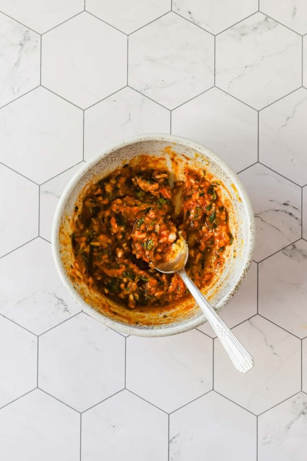 A ceramic bowl containing a chunky, orange-red mixture with herbs, and a metal spoon, placed on a white hexagonal tile surface.