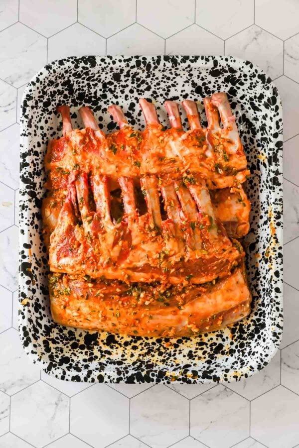 A speckled baking dish containing two racks of marinated lamb ribs coated with herbs and spices, placed on a light, hexagonal tile surface.