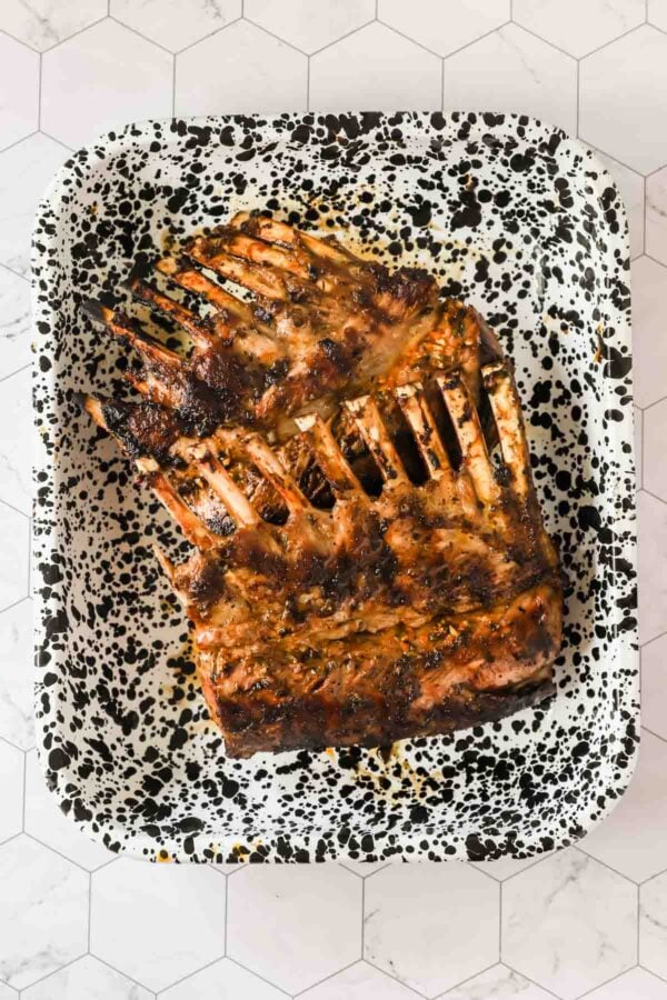 A cooked rack of lamb with seasoning sits in a speckled black and white roasting pan on a white tile surface.