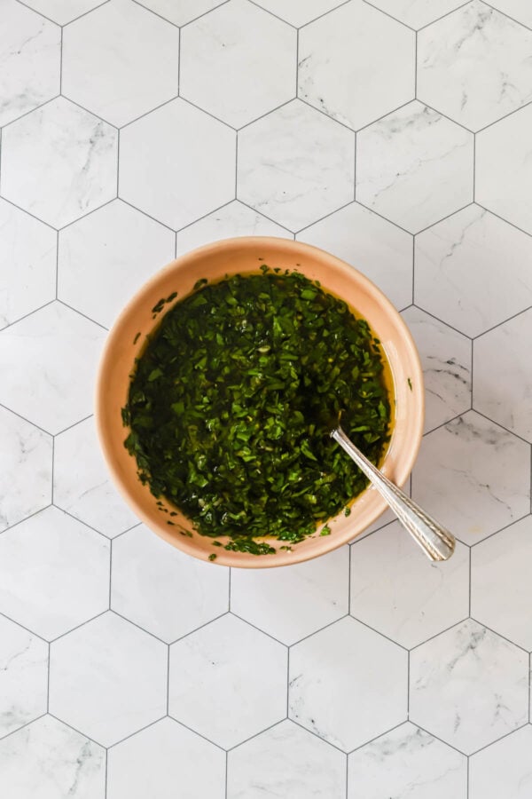 A bowl of chopped green herbs with a spoon, placed on a white hexagonal tile surface.