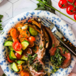 A plate with grilled rack of lamb topped with green sauce, served with a tomato and cucumber salad, placed on a blue and white dish next to fresh parsley and tomatoes.