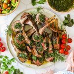 A plate of grilled lamb chops topped with green herb sauce, garnished with fresh rosemary, cherry tomatoes, and served alongside salad bowls.