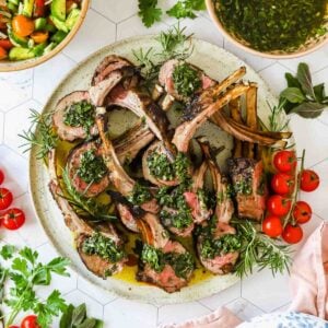A plate of grilled lamb chops topped with green herb sauce, garnished with fresh rosemary, cherry tomatoes, and served alongside salad bowls.