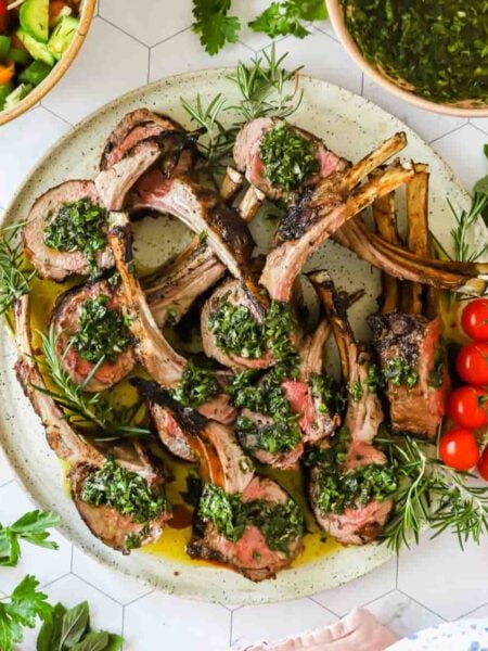 A plate of grilled lamb chops topped with green herb sauce, garnished with fresh rosemary, cherry tomatoes, and served alongside salad bowls.