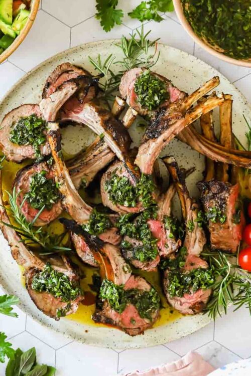 A plate of grilled lamb chops topped with green herb sauce, garnished with fresh rosemary, cherry tomatoes, and served alongside salad bowls.