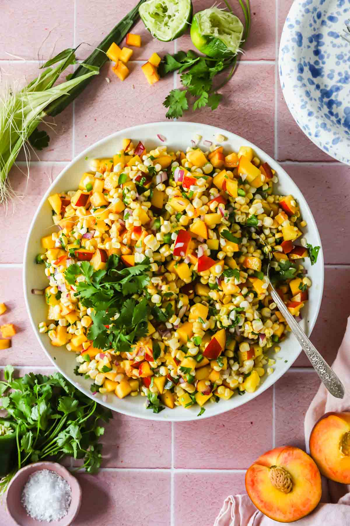 A bowl of corn and peach salad garnished with cilantro, surrounded by fresh herbs, lime halves, and sliced peaches on a pink tiled surface.