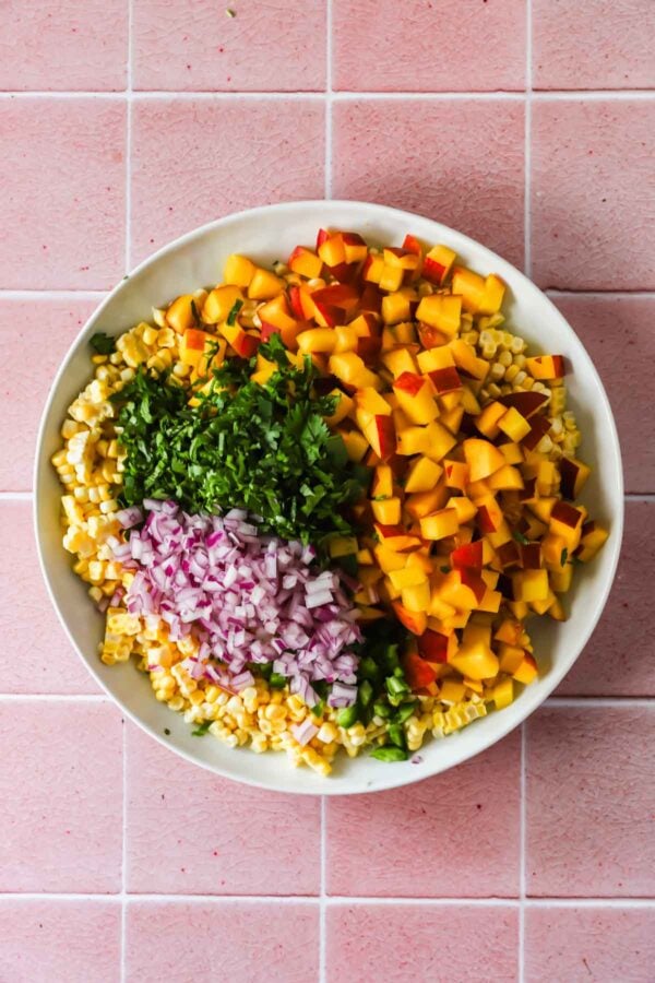 A white bowl filled with chopped peaches, corn, red onion, jalape&ntilde;o, and cilantro sits on a pink tiled surface.