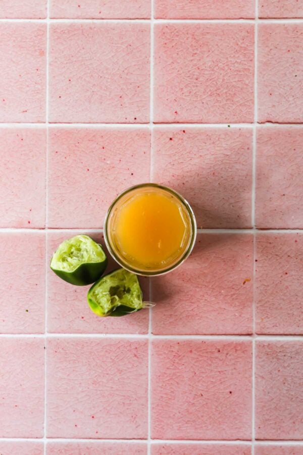 A glass of orange juice and two squeezed lime halves on a pink tiled surface.
