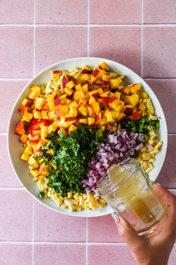 A hand pours dressing from a jar onto a bowl of chopped peaches, corn, red onion, jalape&ntilde;o, and cilantro on a pink tiled surface.