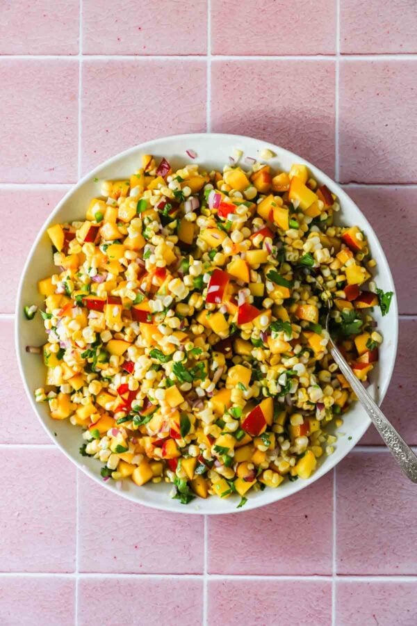 A bowl of corn salad with diced peaches, red onion, cilantro, and bell pepper on a pink tiled surface, with a spoon in the bowl.