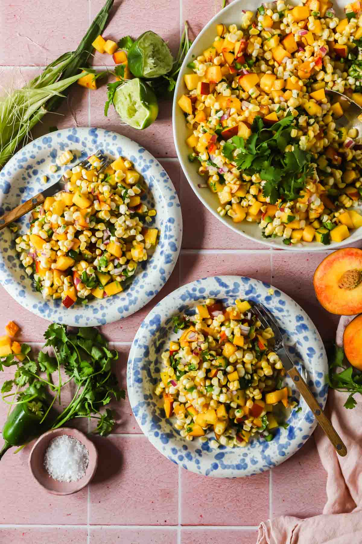 Three plates with corn and peach salad garnished with cilantro are on a pink tile surface, with fresh herbs, limes, a jalape&ntilde;o, and a peach nearby.