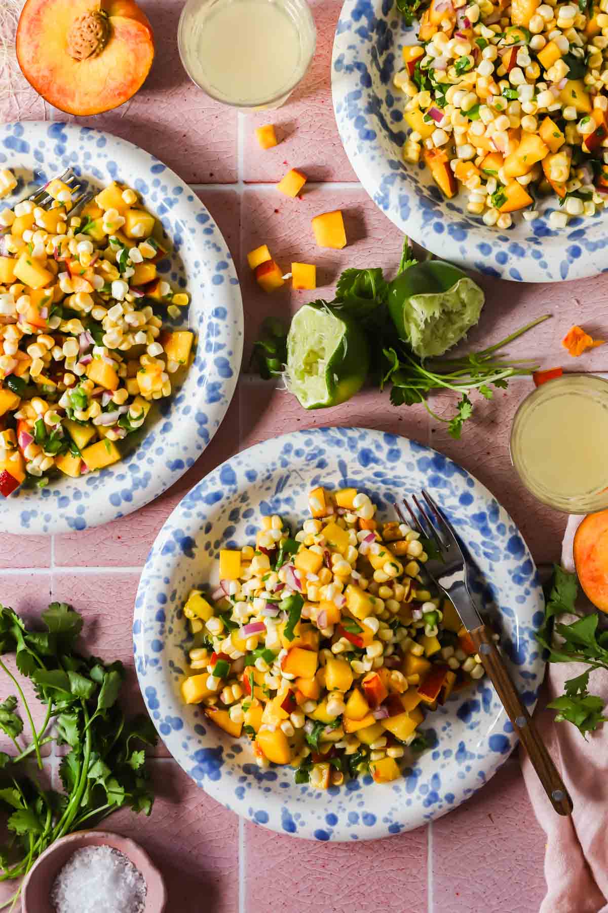 Two blue and white plates filled with corn, diced peaches, and chopped vegetables on a pink tiled surface, with fresh peaches, lime, cilantro, and drinks nearby.