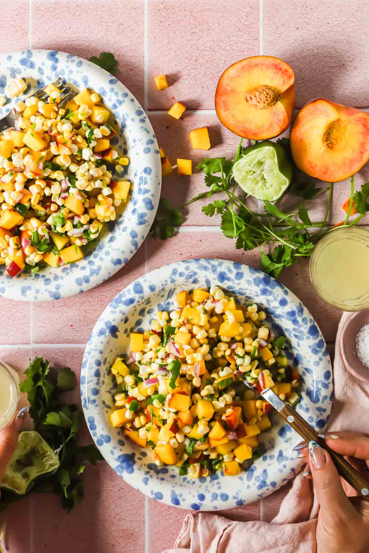 Two blue and white plates filled with corn, diced peaches, and chopped vegetables on a pink tiled surface, with fresh peaches, lime, cilantro, and drinks nearby.