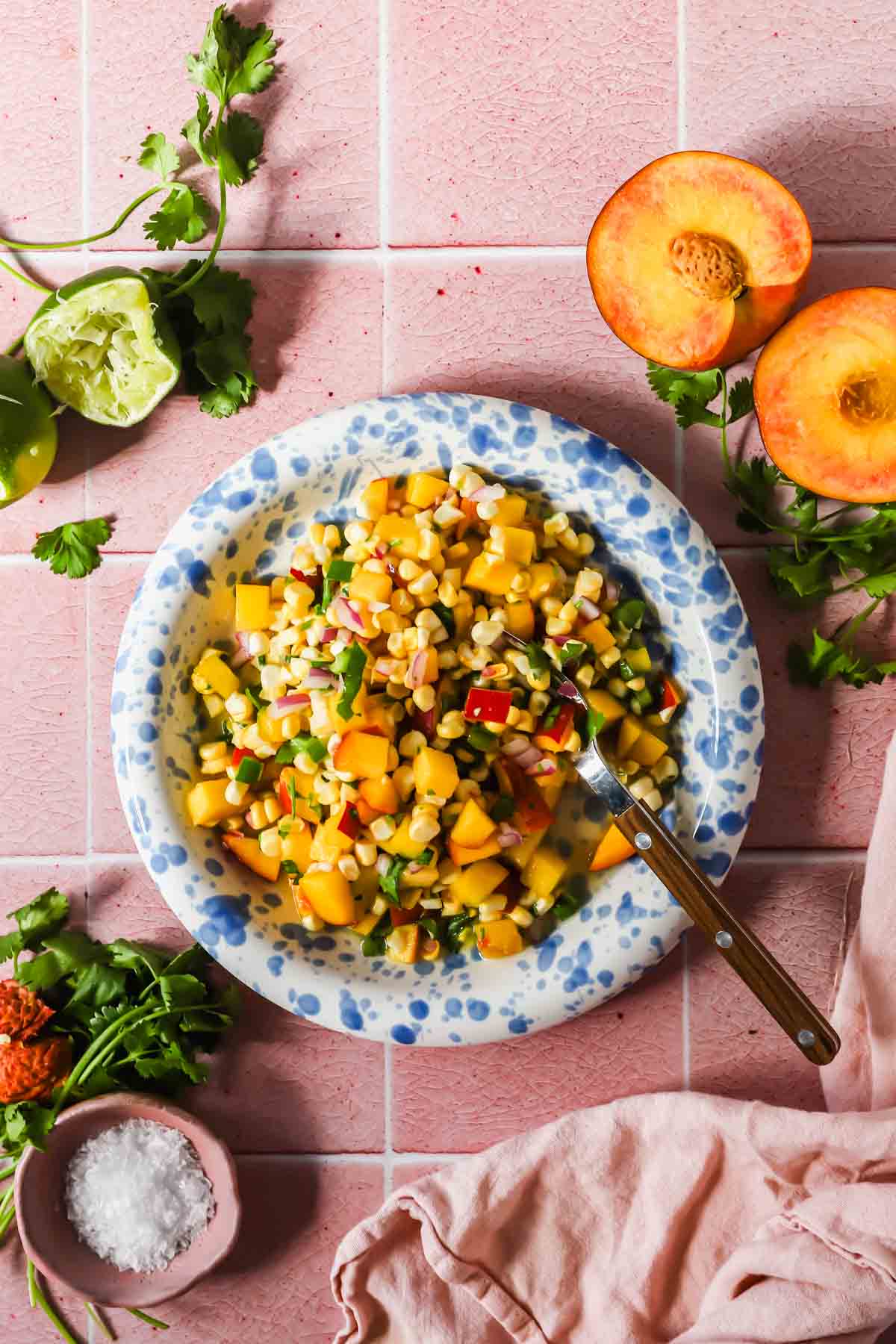 A plate of corn and peach salad with diced vegetables, surrounded by fresh cilantro, lime halves, a halved peach, salt, and a fork on a pink tiled surface.