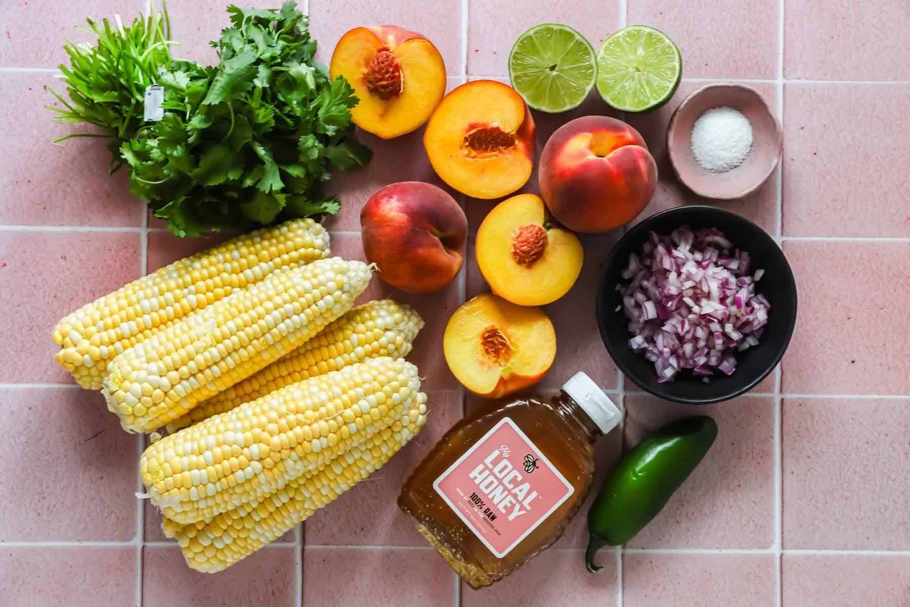Ingredients arranged on a pink tile surface: peaches, corn, cilantro, limes, salt, diced red onion, jalape&ntilde;o, and a bottle of local honey.
