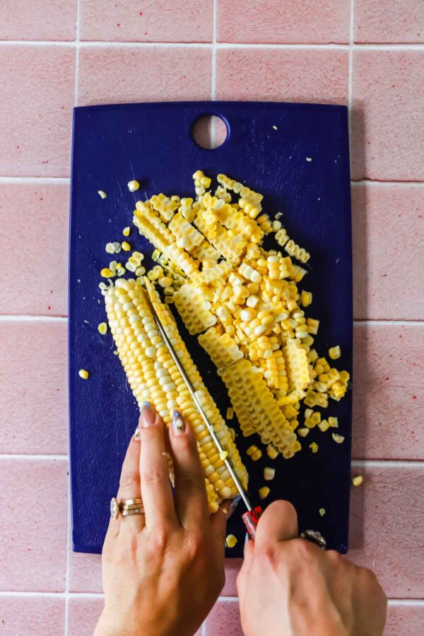 A person cuts corn kernels off a cob on a dark blue cutting board placed on a pink tiled surface.