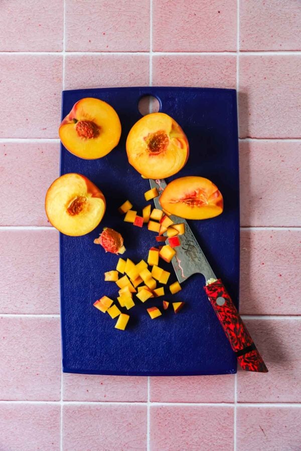 Halved peaches, peach cubes, and a knife with a red handle on a blue cutting board, set on a pink tiled surface.