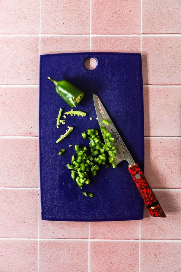 A chopped jalape&ntilde;o pepper and a knife with a red handle on a blue cutting board, placed on a pink tiled surface.