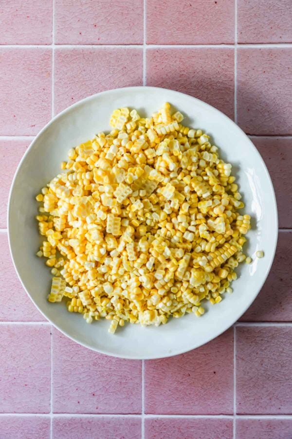 A white plate filled with cut kernels of yellow and white corn sits on a pink tiled surface.